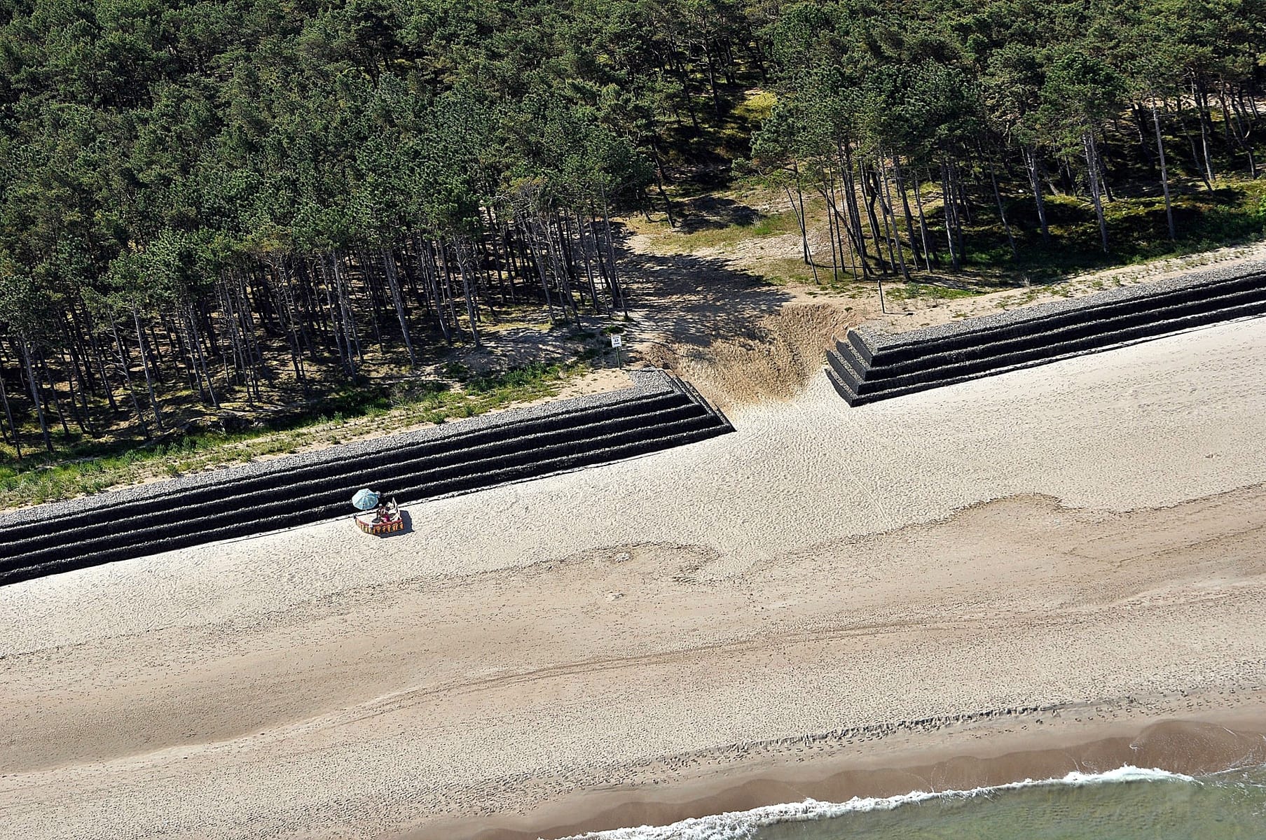 Zdjęcie z lotu ptaka przedstawiające opaskę z koszy gabionowych, zbudowaną pomiędzy plażą a lasem.
