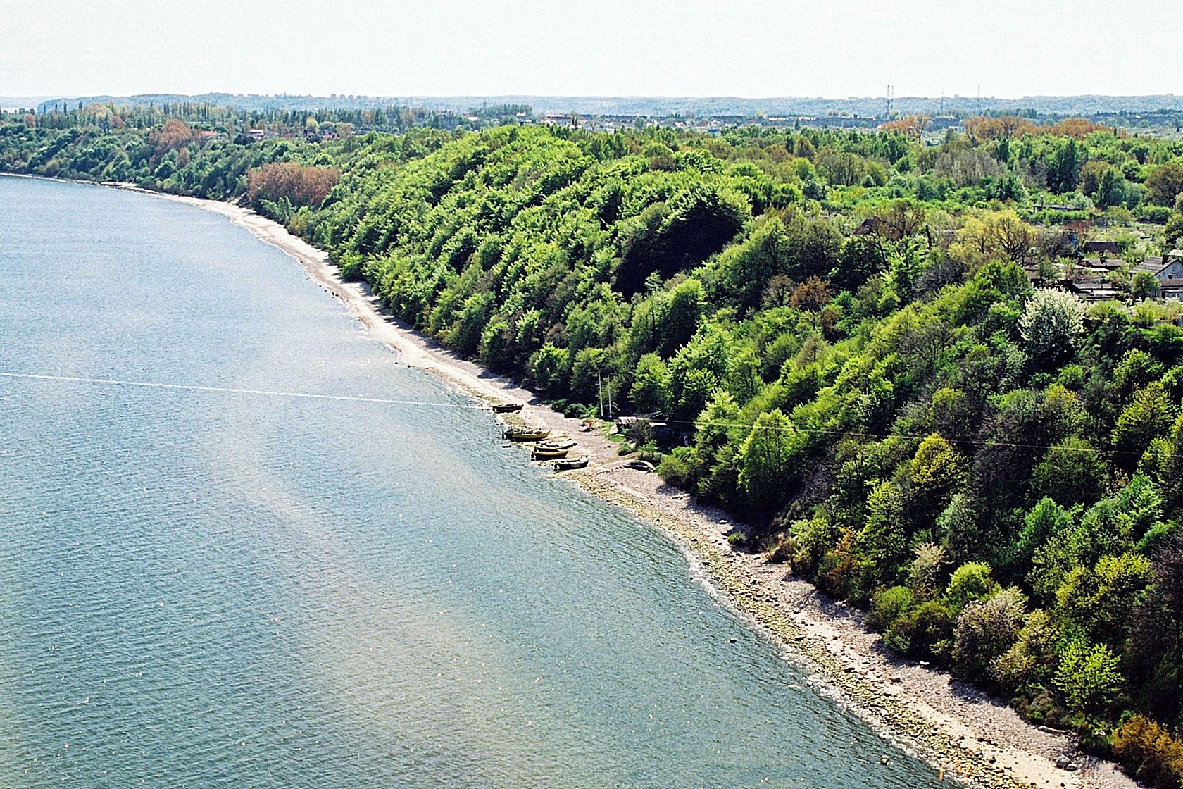 Zdjęcie z lotu ptaka ukazujące zbocze klifowe gęsto porośnięte roślinnością wysoką, wąską kamienistą plażę oraz przystań rybacką.