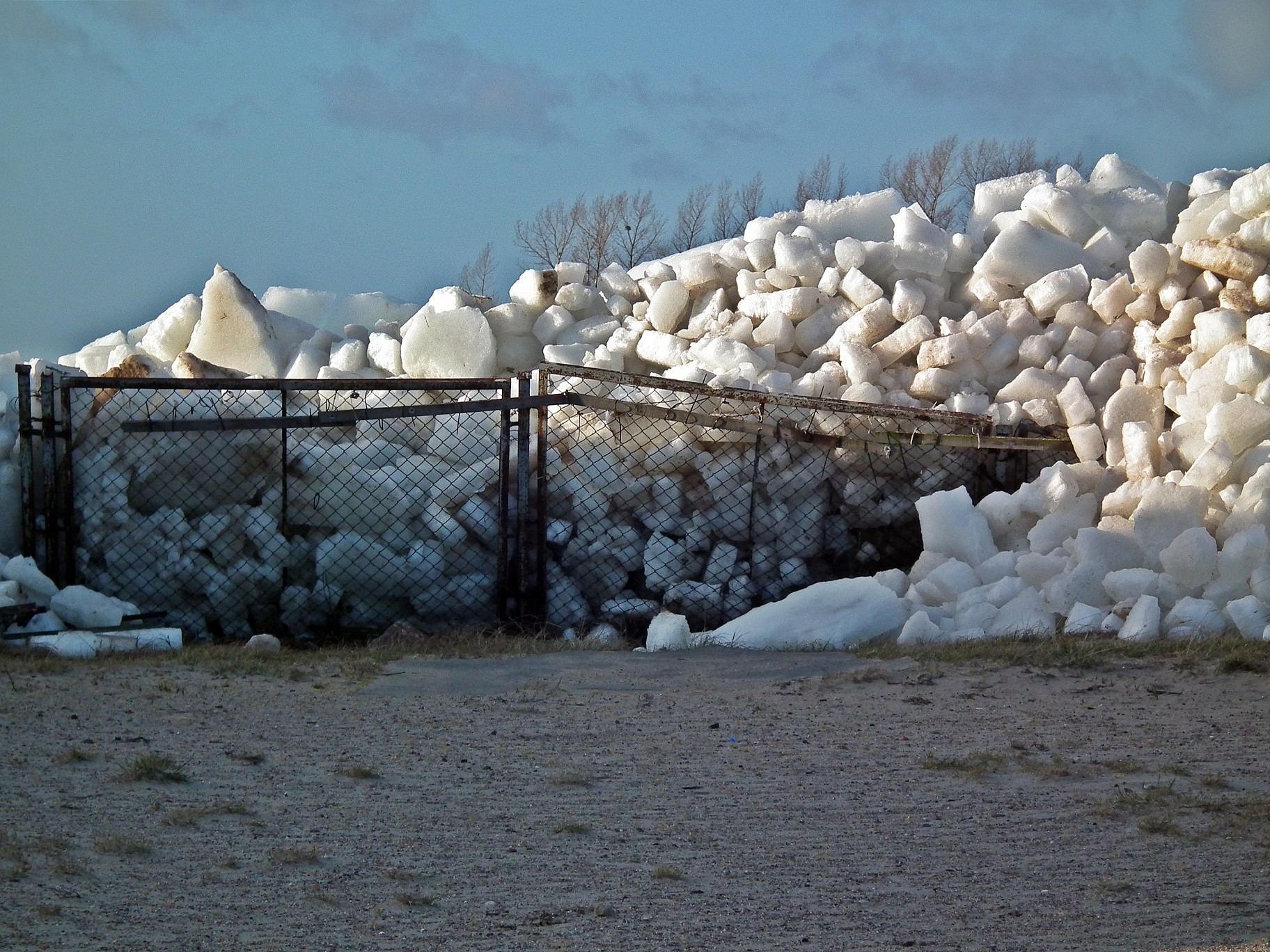 Zdjęcie przedstawiające ogromną ilość kry lodowej, wepchniętej przez fale na ląd w Chałupach. Widoczna góra lodu oraz zniszczone ogrodzenie.