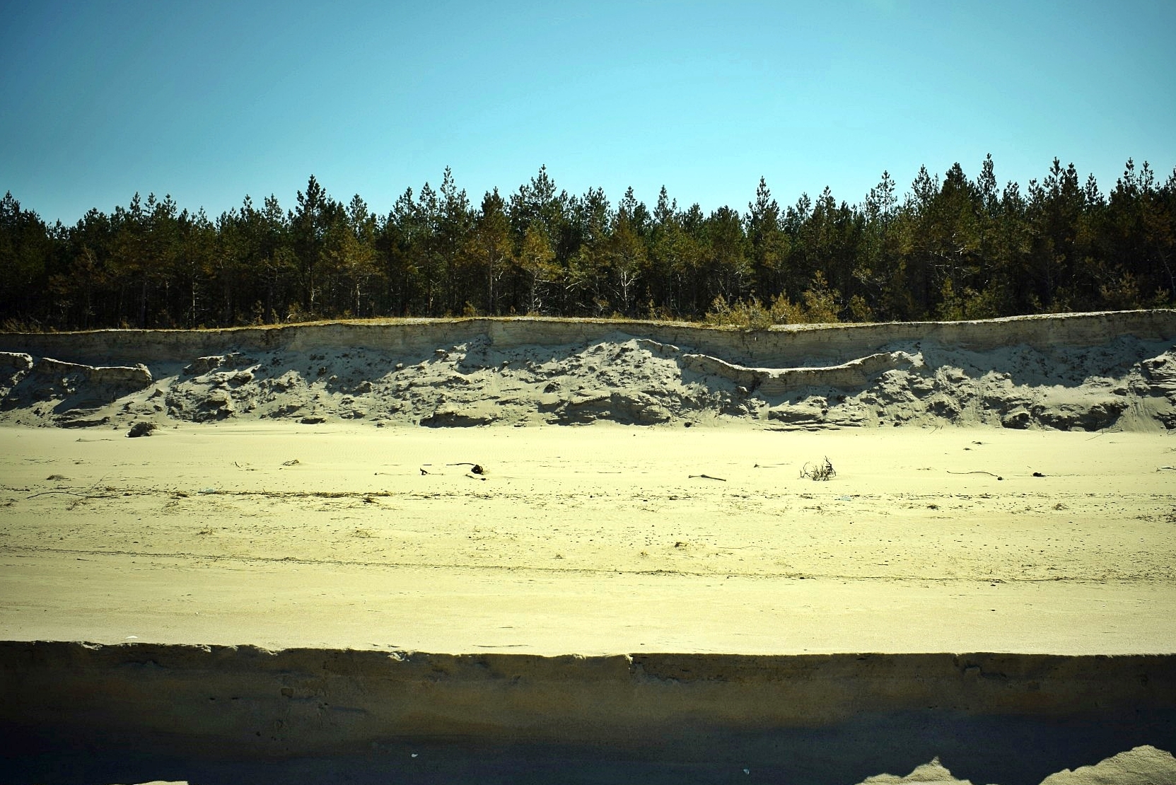 Zdjęcie zrobione z linii brzegu, prostopadle w kierunku lądu, obrazujące stopień na plaży, utworzony przez zmycie części materiału przez fale. Na drugim planie pozostałości wydmy białej po przejściu sztormu.
