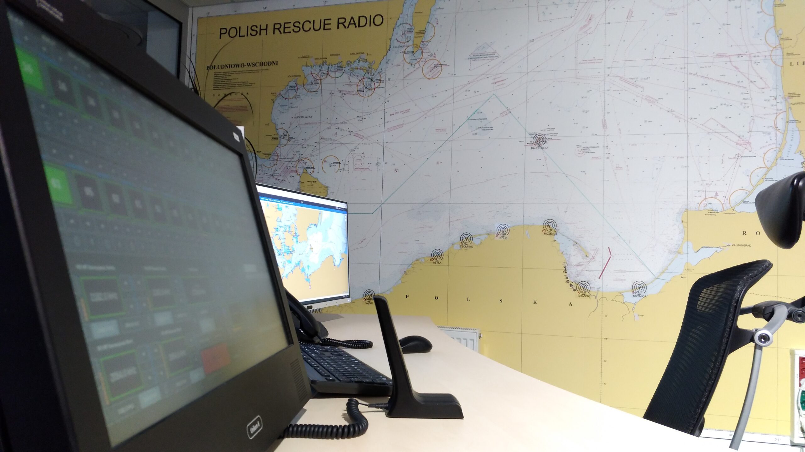 Polish Rescue Radio operator's workplace. On the wall, there is a large navigational map of the Polish coast with the location of radio stations, their frequencies and bands. A fragment of the desk. There are two black computer monitors, a black keyboard, a black telephone and a black microphone on the table top. On the screen of the closer monitor you can see the icons of the individual radio stations allowing you to select stations, frequencies and other functions. The second monitor displays an application for identifying the position of ships - the map of the Baltic Sea