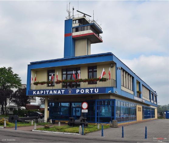 The building of the Harbor Master's Office in Gdynia - yellow and blue, long, glazed, two-story building with a high, blue and yellow tower with a port traffic control room and radar scanners. On the gable wall - on the ground floor, a setback glass entrance to the building. Above the entrance there is a balcony decorated with white and red flags and greenery